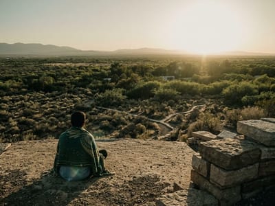 Person sitting alone on a rock ledge facing open desert scrubland at sunset