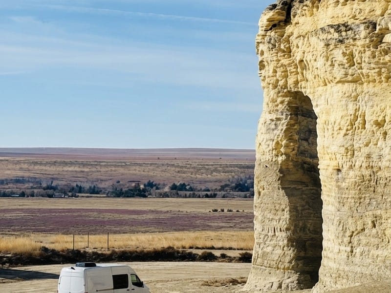 A white camper van parked near a large limestone rock formation with an arched opening, overlooking an expansive prairie under a blue sky.