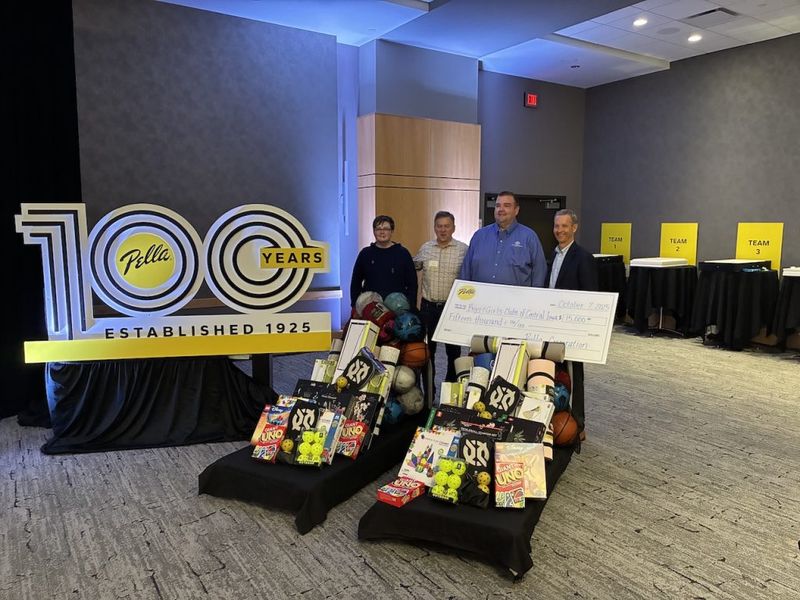 Four people holding a large ceremonial check at a Pella 100th anniversary team-building event, with prize tables stacked with games and sports equipment in the foreground