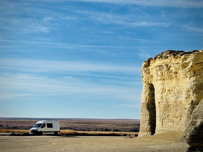 White camper van parked beside a tall chalk rock formation on an open plain under a blue sky