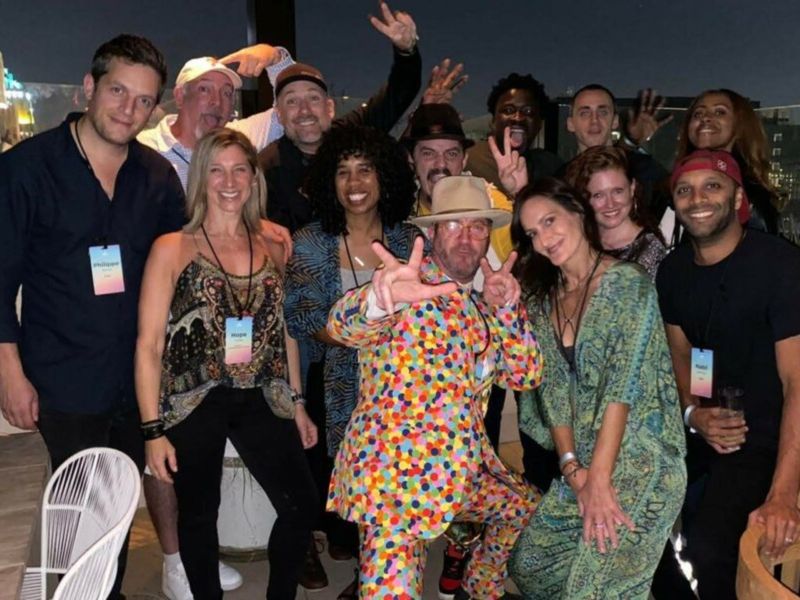 Group of cityHUNT participants celebrating on a rooftop at night, with a man in a colorful polka-dot suit and wide-brimmed hat making peace signs at center