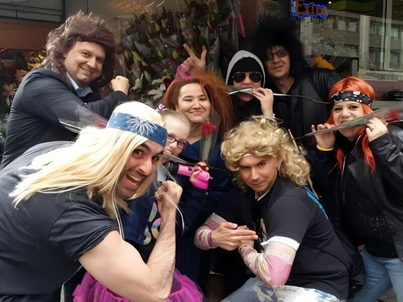 Group of coworkers wearing colorful rock-star wigs and costumes laughing together on a city street during a cityHUNT team-building scavenger hunt