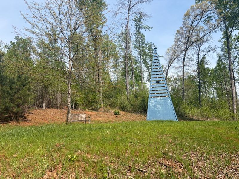 A tall blue pyramid-shaped wooden structure in a grassy woodland clearing, with a bench visible among the trees in the background
